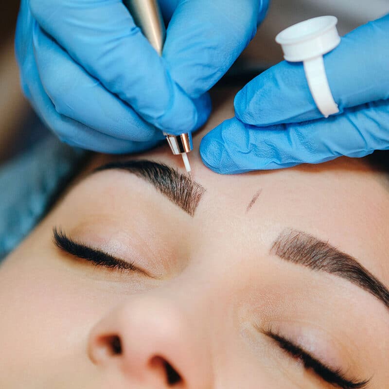 Permanent make-up for eyebrows of beautiful brunette woman in beauty salon. Closeup beautician in gloves doing microblading for eyebrow.
